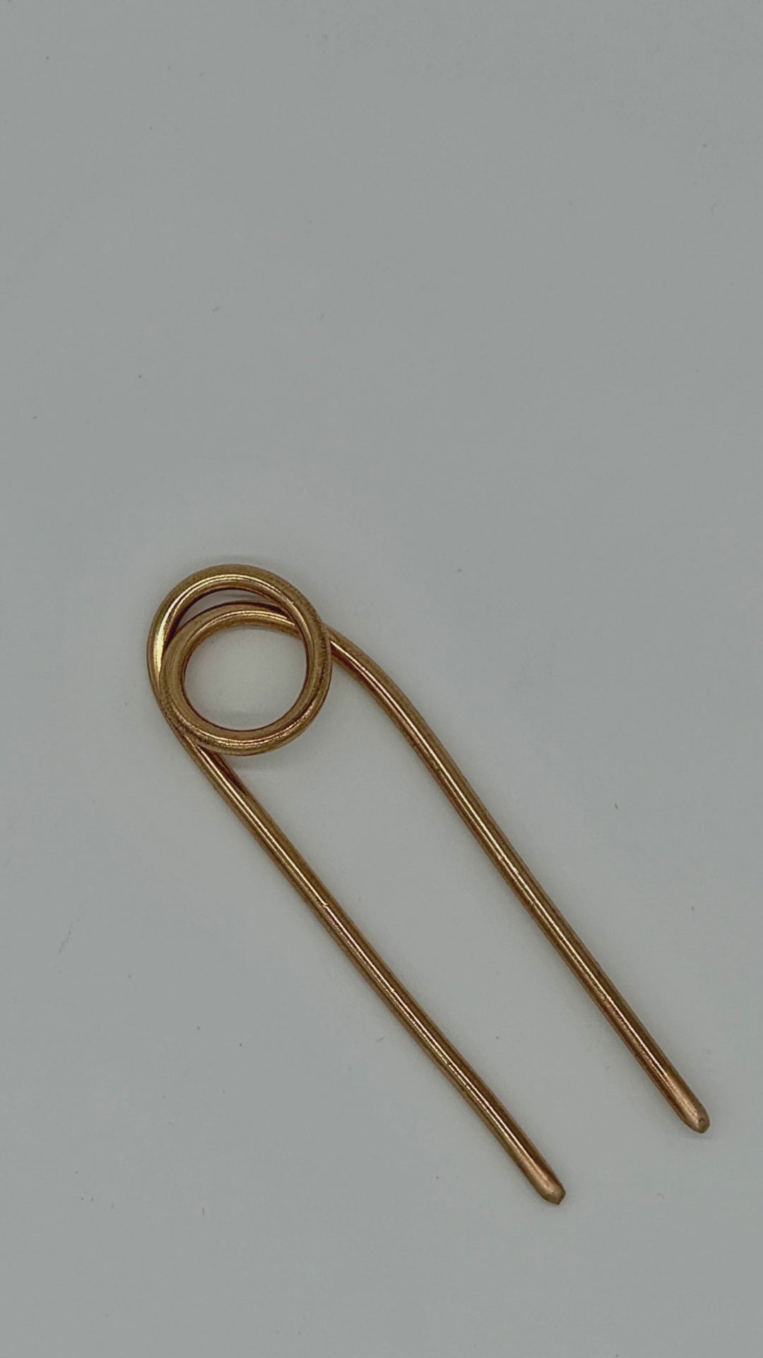 A video of a women demonstrating how to use a brass hairpin. Steps include twist hair into a bun, insert hairpin from the bottom of the bun, then flip the hairpin and push back through the top of the bun.
